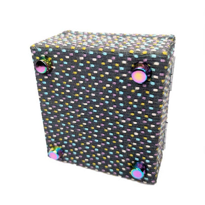 A square dark charcoal woven dice box covered in a dense multicolor dotted pattern of small pastel and bright dots, with four iridescent round metal feet on the bottom, shown on a plain white background.