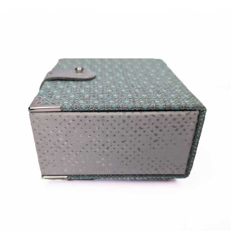 A rectangular dice box with a textured patterned fabric exterior in teal and gray, featuring a small leather strap closure with a metal snap on the lid, and silver-toned metal corner protectors, shown on a plain white background.