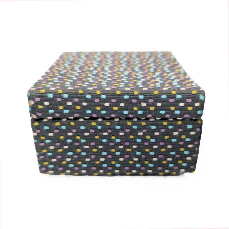 A square fabric-covered box with a lid, featuring a dark charcoal background covered in a dense pattern of small multicolored dots in blue, yellow, purple, and white.